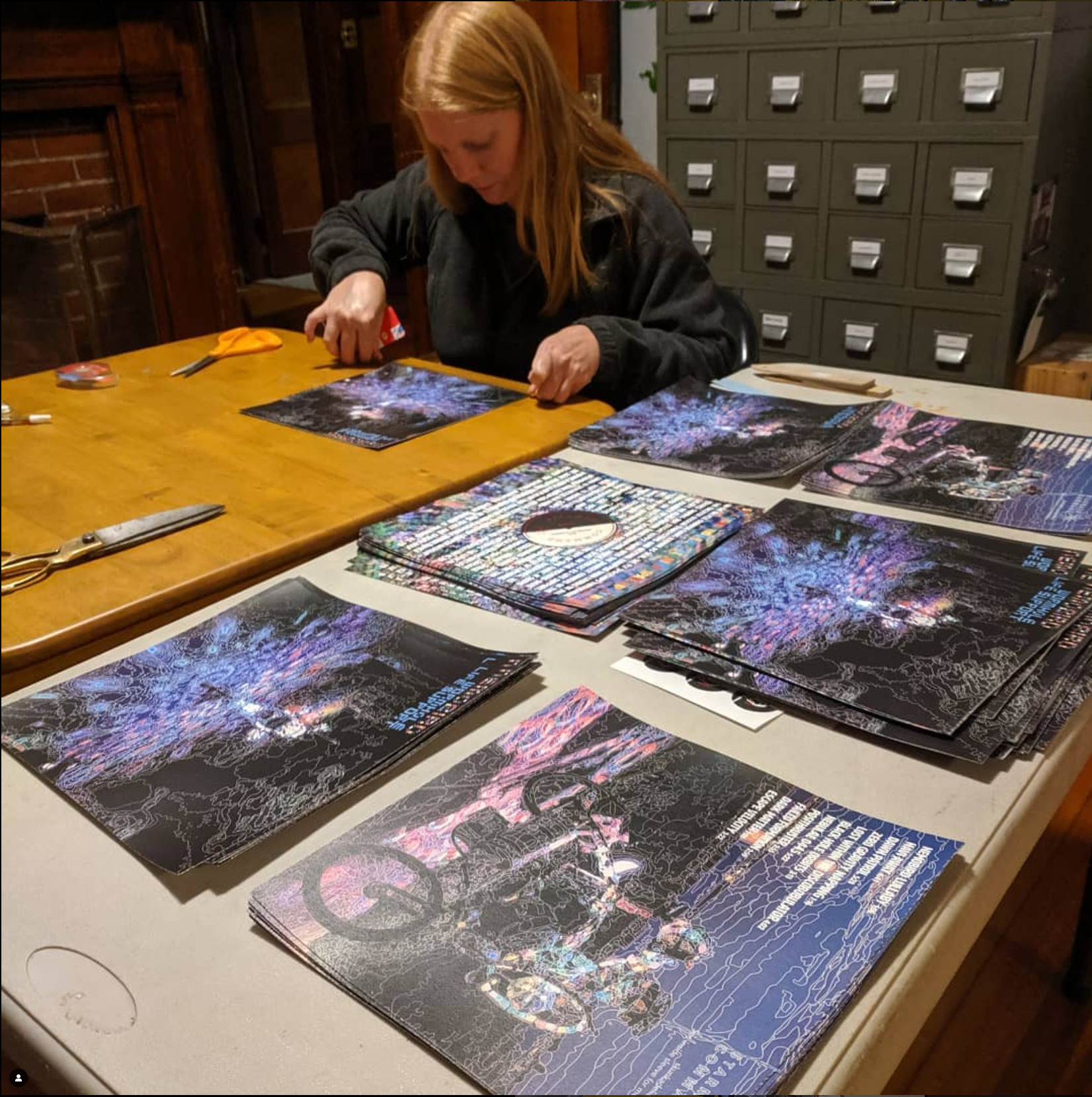 a woman sits at a table assembling stacks of printed paper into a record album sized paper sleeve with a paper insert printed to look like a vinyl album in its sleeve to go inside.