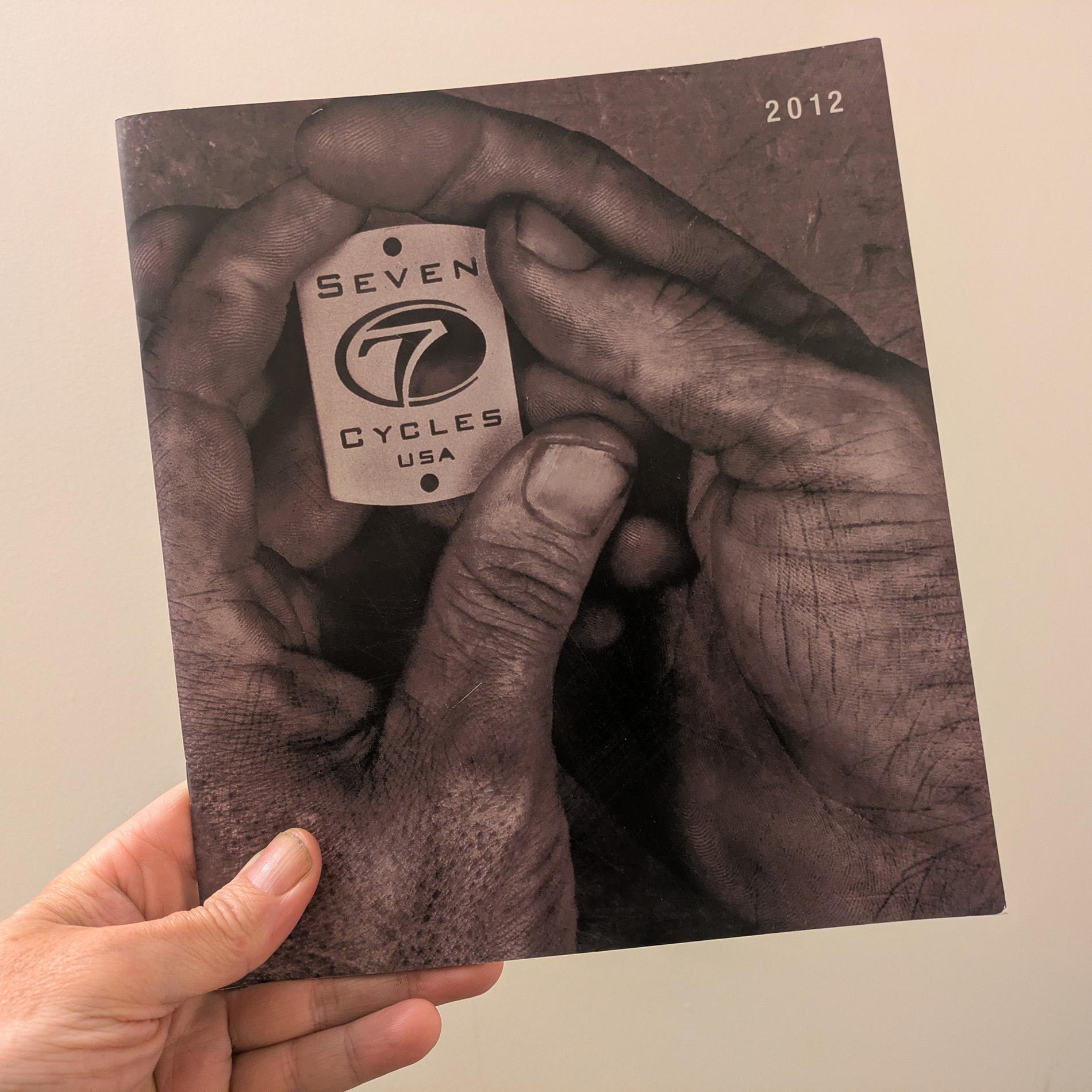 Seven Cycles 2012 Brochure in hand - the cover is a black and white photo of a pair of dirty hands holding an unbent laser cut headbadge that reads 'Seven Cycles USA'