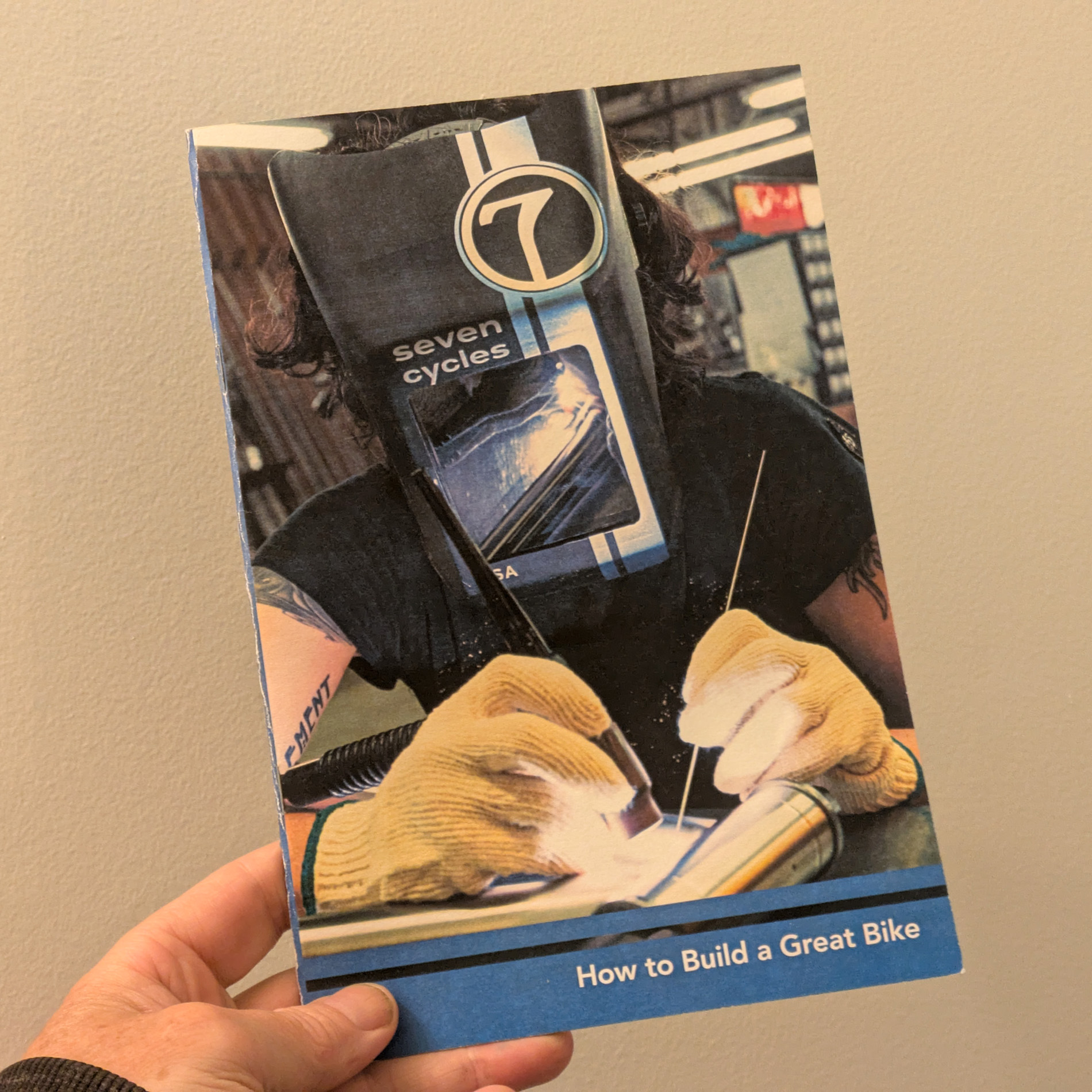Love to Ride Book in hand. The cover is a closeup of a man's hands filing a laser cut titanium lug on a workbench cluttered with titanium tubes, files, and a dial caliper.