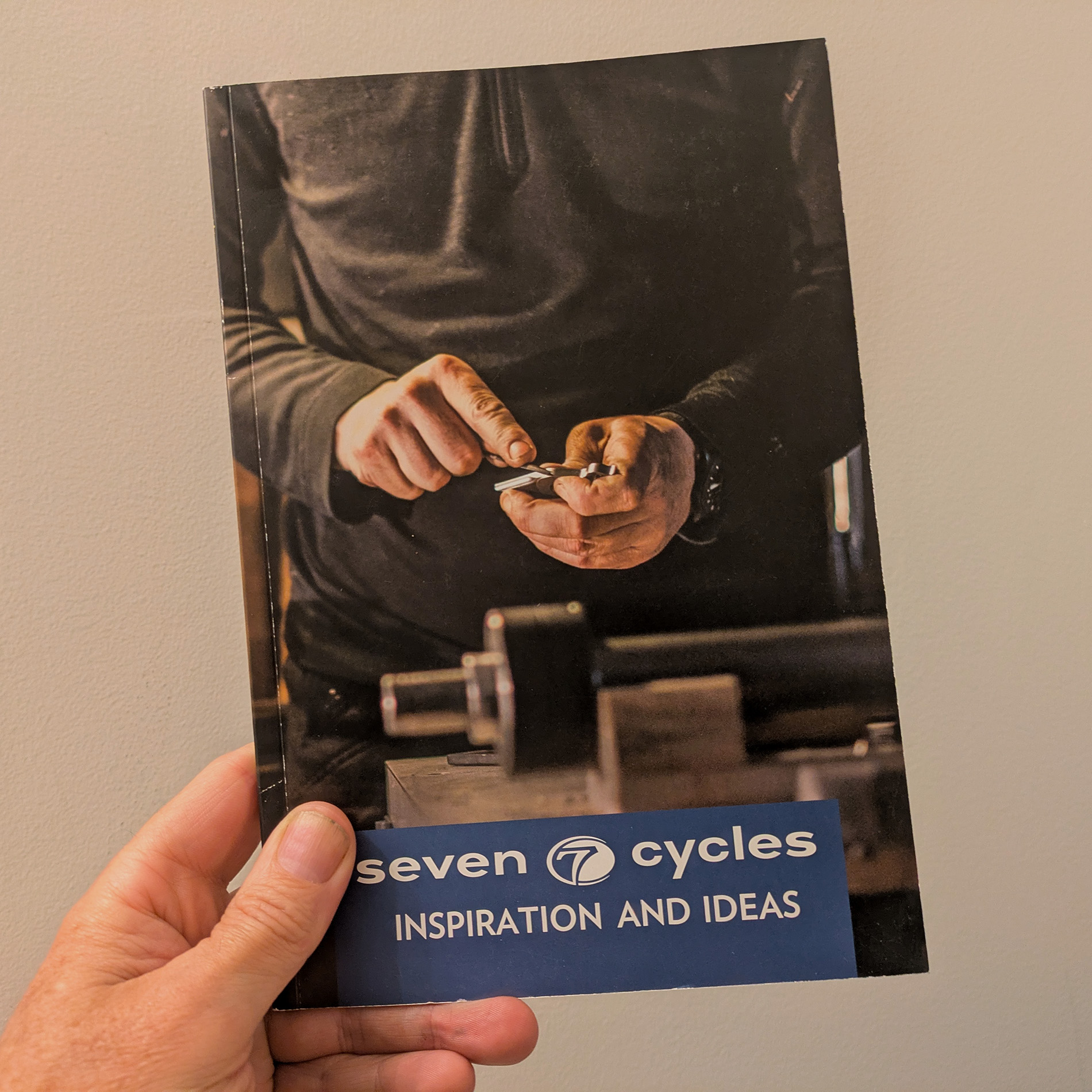 Love to Ride Book in hand. The cover is a closeup of a man's hands filing a laser cut titanium lug on a workbench cluttered with titanium tubes, files, and a dial caliper.
