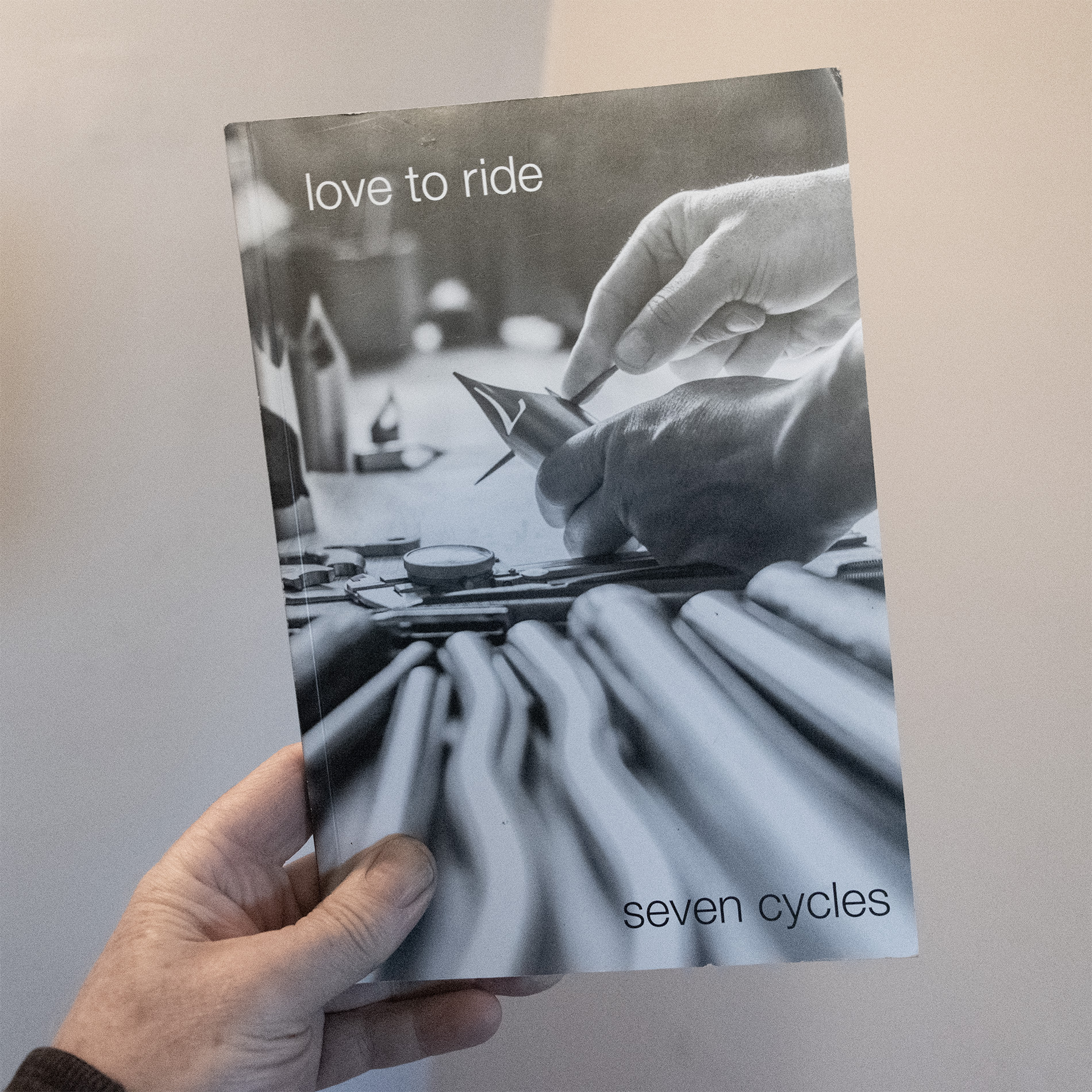 Love to Ride Book in hand. The cover is a closeup of a man's hands filing a laser cut titanium lug on a workbench cluttered with titanium tubes, files, and a dial caliper.