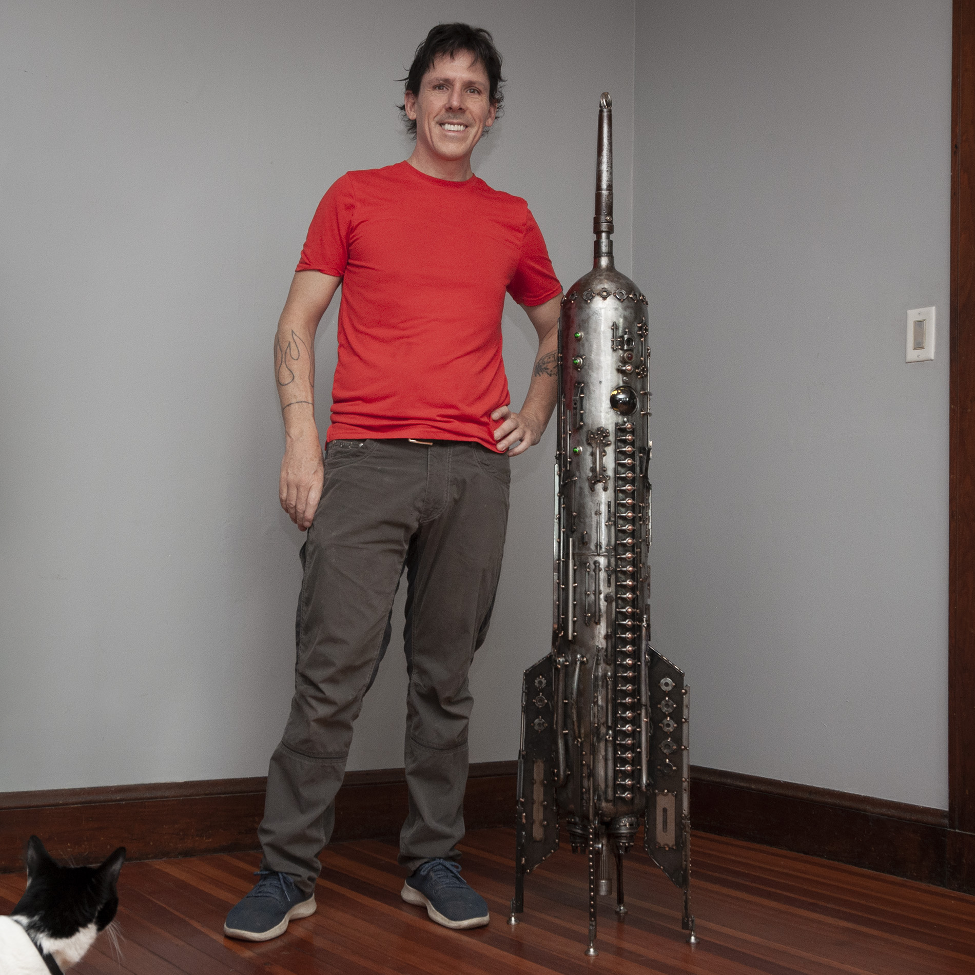 a proud sculptor standing next to an ornate rocketship sculpture made from repurposed scuba tanks