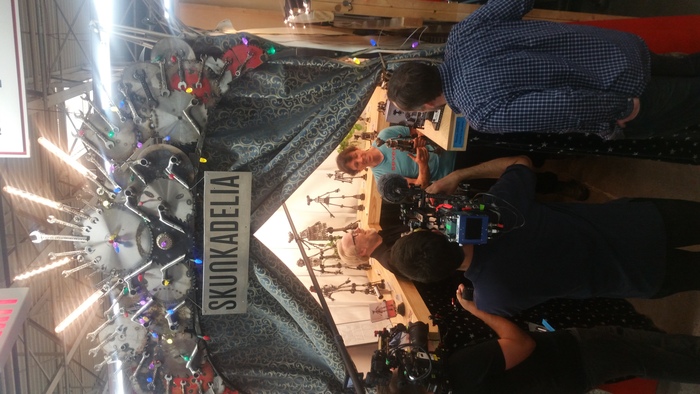 Adam Savage and his film crew crowd into a tiny gallery space where a man holds a small metal sculpture of a robot amongst many.