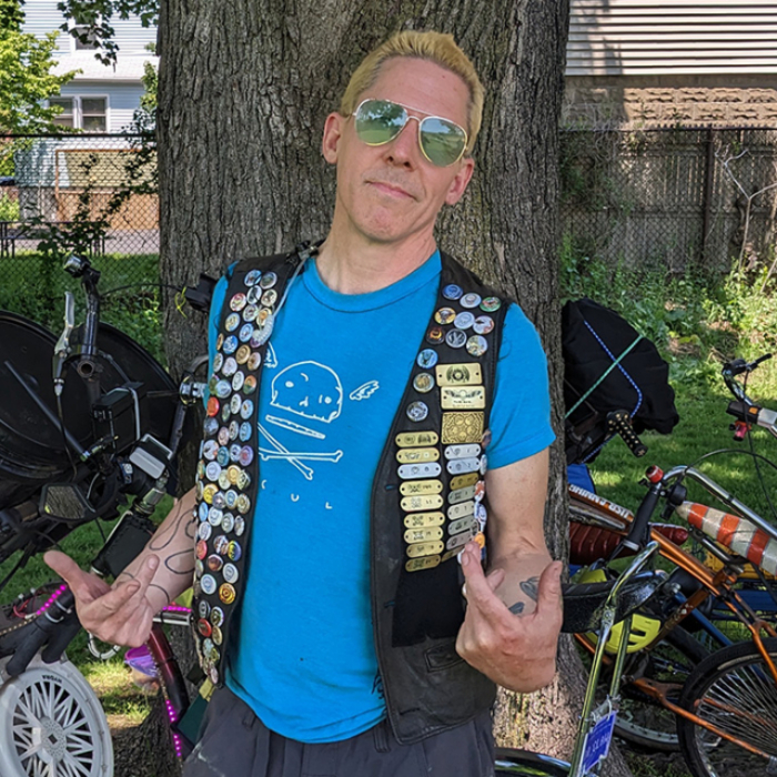 A smirking blonde man in blue aviator sunglasses shows off his vest covered in one inch buttons, each with a different design on them.