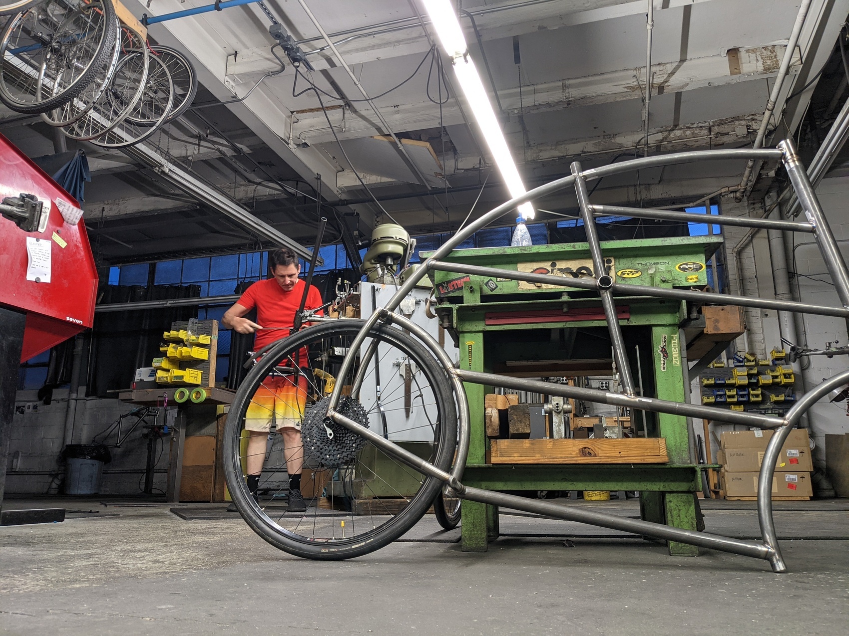 a half-built bicycle frame leans against a surface place with a machinist filing a steel tube in the background