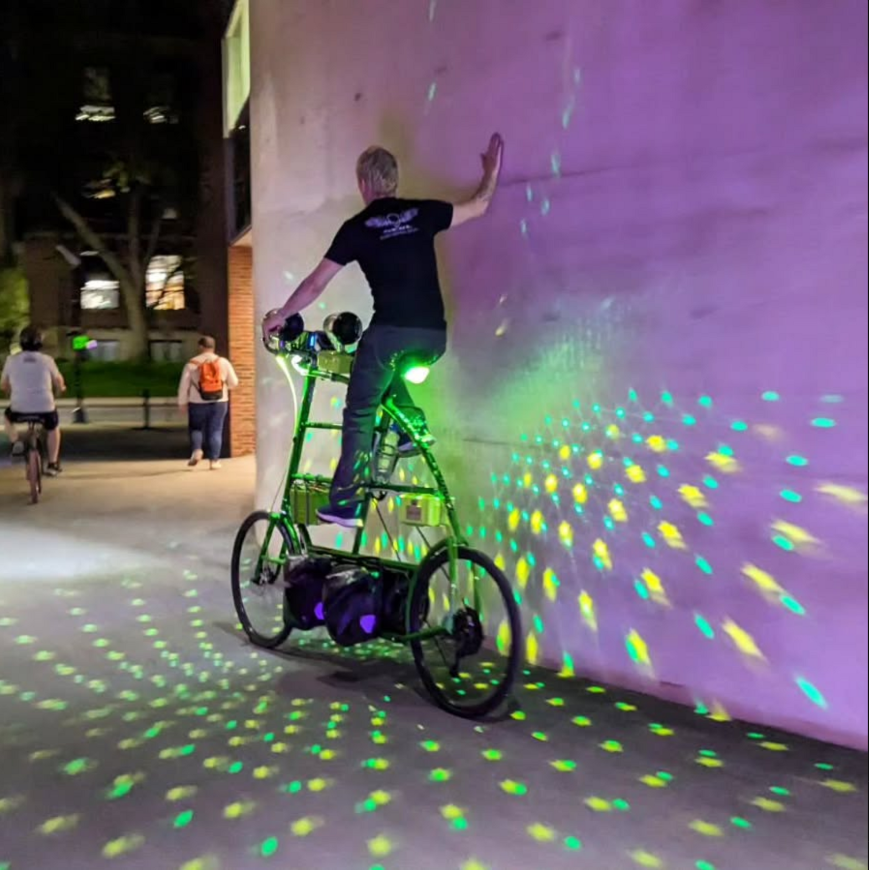 an expertly crafted tallbike with green lights projected all around it, rides alongside a wall in the city at night.