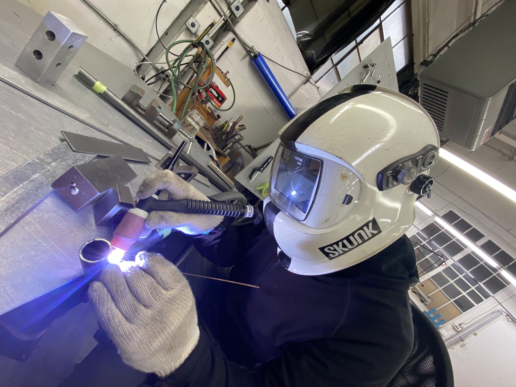 long steel tubes are precisely tig welded together in a clean shop with a big aluminum weld table. The helmet reads 'skunk' on the side